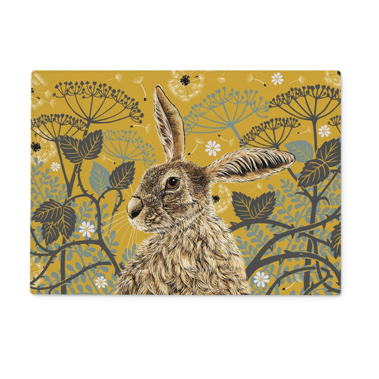 This worktop saver design features a hare portrait with one floppy ear, around the hare are stylised brambles, leaves, cow parsley and dandelion puffs, background colour is a mustard yellow.